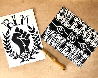 Puede incluir: Dos impresiones de linóleo en blanco y negro sobre papel blanco. La primera impresión presenta un puño en alto con una corona y las letras BLM. La segunda impresión presenta el texto "Silence is Violence" en una fuente audaz y estilizada. También se muestra una herramienta de talla de madera.