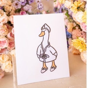 May include: A white card featuring a cartoon illustration of a goose wearing a white bathrobe and slippers. The goose has a yellow beak and orange feet.