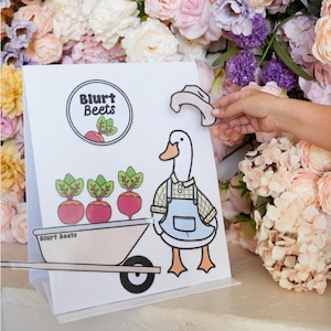 May include: A white sign featuring a cartoon goose wearing a blue overall and a white hat. The goose is pushing a wheelbarrow containing three red beets. The sign reads "Blurt Beets" in a black circle.