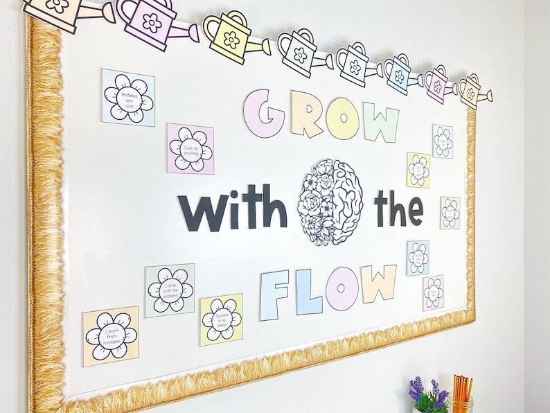 May include: A bulletin board with a gold fringe border and a colourful banner of watering cans. The board has the words "GROW with the FLOW" in large letters. There are also several small flower-shaped cards with positive affirmations written on them.