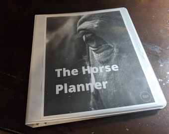 May include: A white binder with a black and white photo of a horse's eye on the cover. The text "The Horse Planner" is printed in white on the cover. The text "THP" is printed in gold on the cover.
