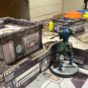Puede incluir: Una figura en miniatura de un soldado futurista, pintada en tonos verdes, azules y plateados, se encuentra sobre una base circular. La figura sostiene un rifle y está situada dentro de un detallado paisaje urbano en miniatura gris y blanco, sugiriendo un juego de mesa.