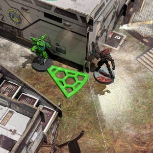 Puede incluir: Una escena de juego de mesa con figuras en miniatura y terreno. Una pieza geométrica verde está cerca de una figura de robot verde y una figura roja y negra. El fondo incluye una fachada de edificio y una superficie estampada.
