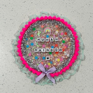 Könnte beinhalten: Ein rosa und silberner, mit Glitzer gefüllter Kreis mit einer weißen Spitzenborte. Der Kreis enthält einen Regenbogen und die Worte "HUMAN RAINBOW", die mit weißen Buchstaben-Kacheln ausgeschrieben sind. Ein kleines rosa Herz befindet sich in der Mitte des Kreises.