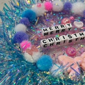 May include: A sparkly blue Christmas wreath with white, pink, and blue pom-poms, a pink bow, and white letter beads spelling "Merry Christmas".