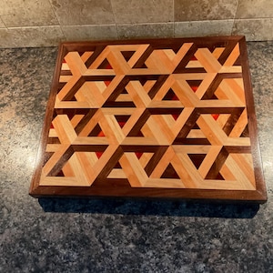 Puede incluir: Tabla de cortar cuadrada de madera con un patrón geométrico. El diseño presenta formas entrelazadas en tonos de madera clara y oscura, con detalles en rojo. La tabla tiene un borde marrón oscuro.