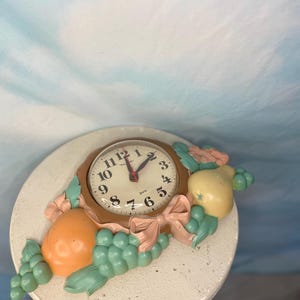 May include: A brown and white quartz clock with a fruit motif. The clock face has black numbers and hands. The fruit motif includes a pear, an orange, and grapes. The clock is attached to a white surface.