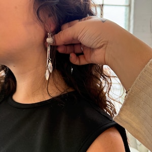 Pode incluir: Brincos pendurados prateados com uma bola texturizada e pingentes em forma de folha. Os brincos são longos e finos, com um acabamento polido. A modelo está vestindo um top preto sem mangas.