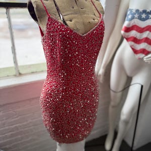 May include: A red, beaded cocktail dress with a V-neck and spaghetti straps. The dress is displayed on a vintage dress form. The dress is covered in silver sequins and has a fitted silhouette.