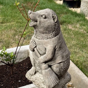 May include: A gray stone garden statue of a hedgehog holding a broom. The hedgehog is wearing a striped shirt and is sitting on a rock.