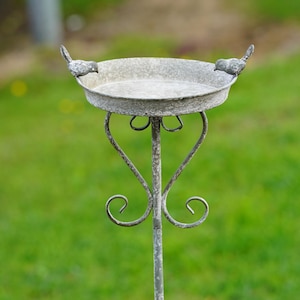 May include: A weathered metal bird feeder with a shallow dish and decorative bird accents. The feeder is supported by a slender, ornate stand with heart-shaped and curled metalwork. The background is a blurred green lawn.