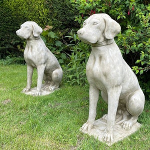 May include: Two grey stone dog statues, one sitting and one standing, are placed on a green grassy lawn. The statues are detailed and realistic, with the dogs' features clearly visible.