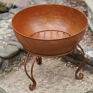May include: A round, bowl-shaped fire pit with a rusted, weathered orange-brown finish. The metal basin features a built-in grate at the bottom and is supported by three curved, scroll-style legs that provide a stable base for the structure.