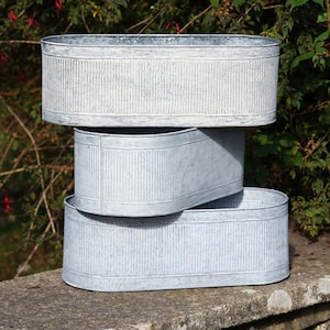 Op de afbeelding: Drie gestapelde, ovale plantenbakken van gegalvaniseerd metaal. De plantenbakken hebben een geribbelde textuur en een verweerde, grijze afwerking. De plantenbakken zijn van verschillende afmetingen en zijn geplaatst op een stenen oppervlak, met groen op de achtergrond.