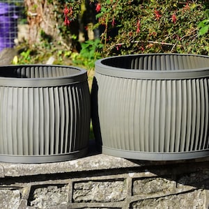 May include: Two olive-green, barrel-shaped planters with vertical ribbed detailing. The planters sit on a stone wall, with a purple bin and greenery in the background. The planters have a rounded top and base.
