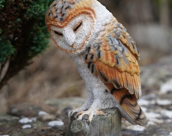 May include: A detailed figurine of a barn owl perched on a tree stump. The owl has closed eyes and is colored in shades of brown, white, and grey. The sculpture is set outdoors with a blurred background of greenery and natural elements.