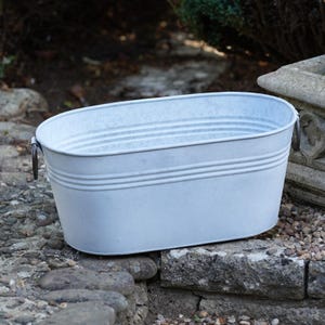 May include: An oval-shaped galvanized metal tub with a smooth, light gray finish. The basin features horizontal ribbed detailing along the upper sides and two dark metal ring handles attached to the left and right ends of the container.