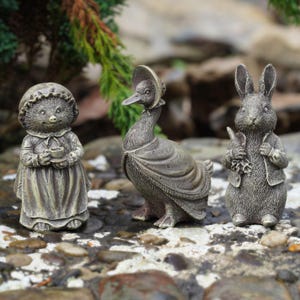 Pode incluir: Três estátuas de jardim detalhadas: um ouriço segurando uma xícara, um pato com chapéu e xale e um coelho segurando uma cenoura e flores. As estátuas têm um acabamento em bronze envelhecido, ideal para decoração ao ar livre. As figuras estão sobre um leito de pequenas pedras.