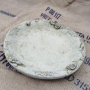May include: A circular, off-white stone bird bath or decorative basin featuring a raised relief pattern of roses and small floral clusters along the outer rim. The surface has a weathered, textured finish with subtle grey and beige colour variations.