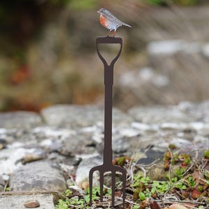 May include: A metal garden fork with a bird perched on top. The fork is dark brown and has a handle with a loop at the top. The bird has a red breast and is facing left. The background is blurred, with a stone wall and greenery.