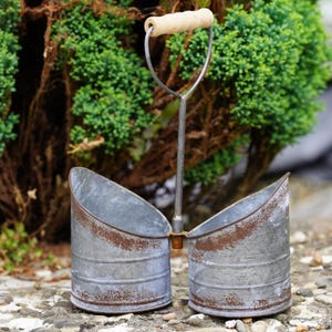 May include: A double-sided galvanised metal caddy featuring two cylindrical containers with a weathered, rusted finish. A central vertical metal rod connects the two sections, topped with a cylindrical light-coloured wooden handle for carrying.