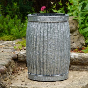 May include: A cylindrical galvanized metal planter featuring a vertical ribbed texture and a smooth, reinforced rim at both the top and bottom. The surface displays a mottled, industrial silver-gray finish with a distinct metallic sheen.