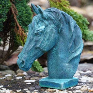 Op de afbeelding: Een verweerde, blauwgroene paardenhoofdsculptuur. De sculptuur heeft gedetailleerde gelaatstrekken en een textuuroppervlak, gemonteerd op een vierkante basis. Het kunstwerk staat buiten, met groen op de achtergrond.