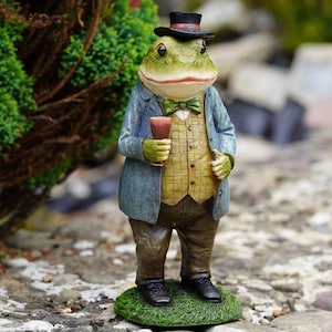 Op de afbeelding: Een staand kikkerbeeldje met een zwarte hoge hoed, een blauwe blazer, een geel geruit vest, een groene vlinderdas en een bruine broek. De kikker houdt een kleine rode beker vast en staat op een gestructureerde groene basis van kunstgras.