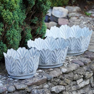 May include: A set of three round, silver-toned metal planters with a scalloped edge design resembling overlapping leaves. Each planter features a textured, weathered finish and a circular base, arranged in a staggered formation from smallest to largest.