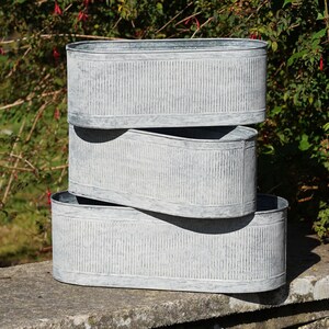 Puede incluir: Tres jardineras ovaladas de metal galvanizado apiladas. Las jardineras tienen una textura acanalada y un acabado gris desgastado. Están sobre un muro de piedra con follaje verde en el fondo. Cada jardinera mide aproximadamente 40 cm de largo.