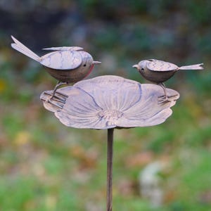 May include: A metal bird bath with two decorative birds perched on a flower-shaped dish. The birds have detailed wings and are made of a weathered metal. The bird bath is on a slender metal pole, perfect for a garden.
