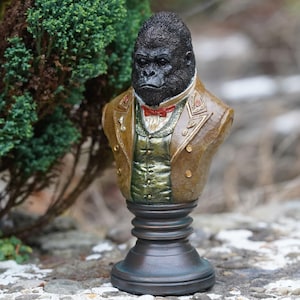 May include: A bust of a gorilla wearing a brown jacket with gold trim, a green vest, and a red bow tie. The gorilla has a serious expression. The bust is mounted on a dark bronze-colored pedestal.