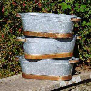 Könnte beinhalten: Drei ovale Pflanzgefäße aus verzinktem Metall, übereinander gestapelt. Jedes Pflanzgefäß hat ein kupferfarbenes Band am Boden und Griffe an den Seiten. Die Pflanzgefäße befinden sich im Freien, mit grünem Laub im Hintergrund.