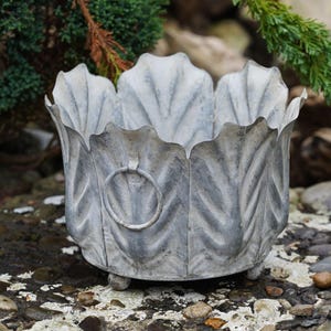Puede incluir: Una maceta de metal gris con un diseño en forma de hoja. La maceta tiene forma redonda con un borde festoneado y un pequeño asa de anillo. Se apoya sobre tres pequeños pies de bola. La maceta es de metal y tiene un acabado envejecido.