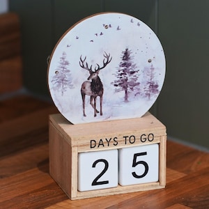 May include: A wooden countdown calendar featuring a circular top panel with a watercolor illustration of a deer in a snowy forest. The base is a rectangular wooden block with the text DAYS TO GO and two white cubes displaying the number 25.