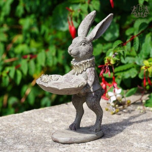 Könnte beinhalten: Eine graue Hasenfigur, die ein dekoratives Tablett hält. Der Hase trägt ein goldenes Halsband und hat eine abgenutzte Oberfläche.