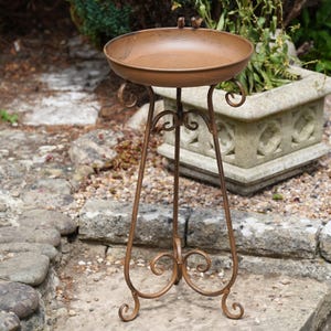 May include: A brown metal bird bath featuring a circular basin with two small bird figurines perched on the rim, supported by a tall, slender stand with three curved legs and decorative scrollwork accents throughout the frame.