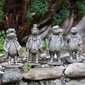Op de afbeelding: Vier grillige tuinbeelden van antropomorfe dieren: een kikker, een beer, een muis en een mol. Elke figuur draagt een vintage pak en hoed, met ingewikkelde details. De beelden zijn in grijstinten en groen, geplaatst op een stenen oppervlak.