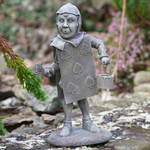 Könnte beinhalten: Bronzefarbene Statue einer Figur aus Alice im Wunderland. Die Figur trägt ein Kartenanzug-Kostüm mit Pik-Symbolen und einen Hut. Sie hält einen kleinen Korb und ein Bündel Zweige und steht auf einem runden Sockel.