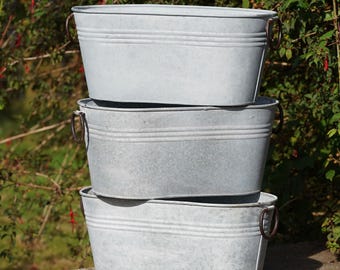 Puede incluir: Tres jardineras ovaladas apiladas de metal galvanizado con asas marrones oscuras. Cada jardinera tiene estrías horizontales cerca de la parte superior y es de color gris claro. Las jardineras están al aire libre, con follaje verde al fondo.