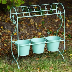 May include: A light mint green metal plant stand featuring a curved, scroll-work frame with an arched top rail and three matching cylindrical pots suspended in a row. The structure is made of thin, painted metal rods with a smooth, matte finish.