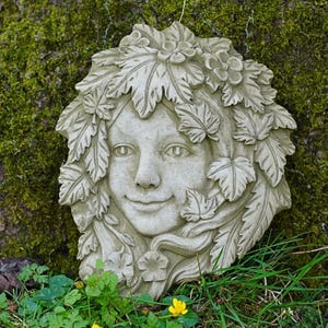 May include: A stone-colored wall plaque featuring a smiling face surrounded by leaves and flowers. The face has detailed features and is framed by a leafy headdress. The plaque is likely for outdoor garden decor.