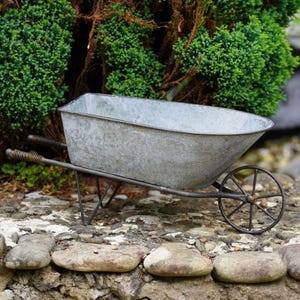 May include: A galvanised metal wheelbarrow planter featuring a rectangular basin with a weathered silver finish, a single thin spoked wheel, and two handles with coiled wire grips, designed for holding plants or decorative garden items.