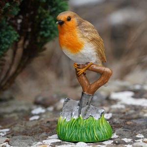May include: A detailed figurine of a robin perched on a miniature shovel. The bird has a bright orange breast, brown wings, and a white belly. The shovel is gray with a wooden handle, set in a green grassy base. A charming garden decoration.