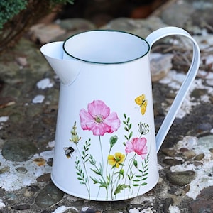 Könnte beinhalten: Weißer Metallkrug mit gebogenem Griff und Ausguss. Der Krug ist mit rosa Mohnblumen, gelben Blumen, grünen Blättern, einem gelben Schmetterling und einer Biene verziert. Der Rand ist grün eingefasst.
