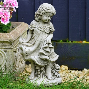 May include: A stone garden statue of a young girl in a flowing dress, looking down at a group of rabbits at her feet. The statue is in a garden setting with green grass and a dark blue fence in the background. The statue is approximately 76 cm tall.
