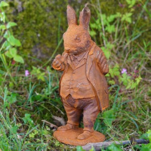 May include: A weathered, rust-colored rabbit statue, dressed in a suit and waistcoat, stands on a small base. The rabbit is posed with one paw raised, as if holding a pocket watch. The garden statue is set against a backdrop of green foliage.