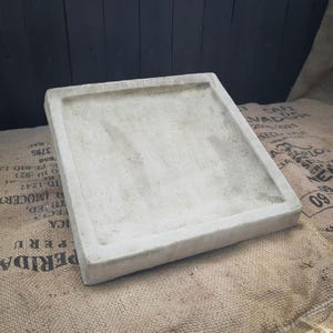 May include: A square, light grey concrete tray with a flat, recessed centre and a raised, thick border. The surface of the tray has a smooth, matte finish with subtle variations in colour and texture throughout the material.