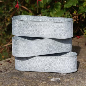 May include: Three stacked, oval-shaped, galvanised metal planters. Each planter features a repeating embossed floral pattern around the sides. The planters are a light grey colour and are sitting on a stone surface.