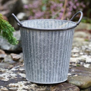 Puede incluir: Un cubo de metal galvanizado gris con dos asas curvas. El cubo tiene detalles verticales acanalados y un borde oscuro. Está sobre una superficie de piedra, con vegetación y flores moradas en el fondo.